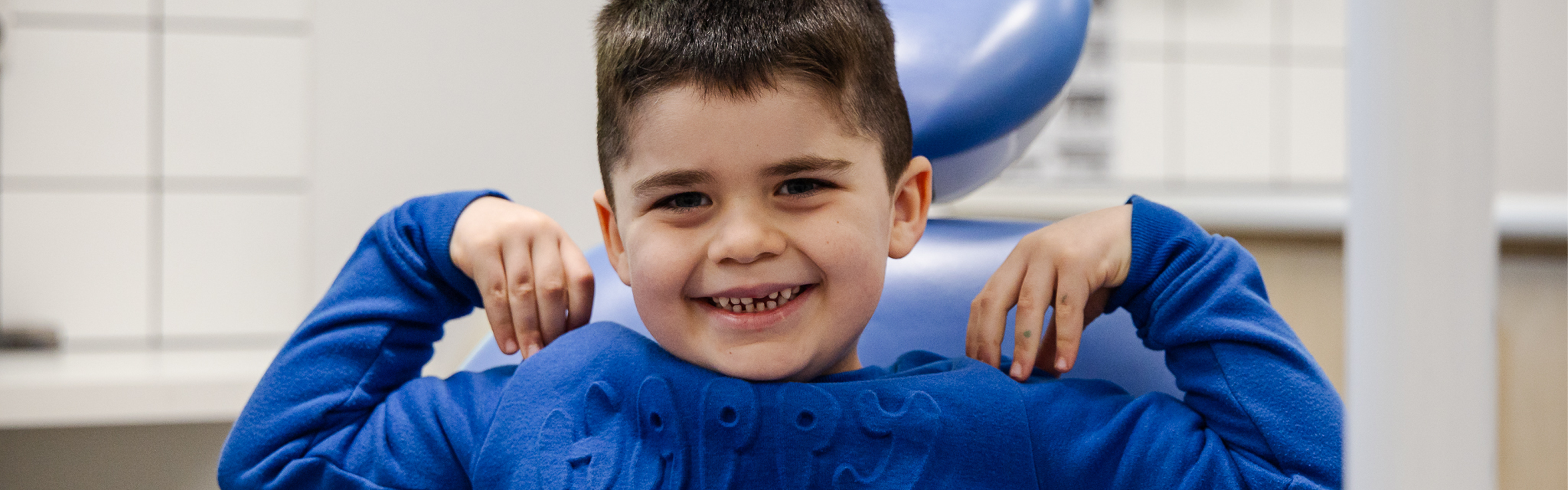 A young boy smiling while sitting in a dental chair and touching his shoulders with his hands