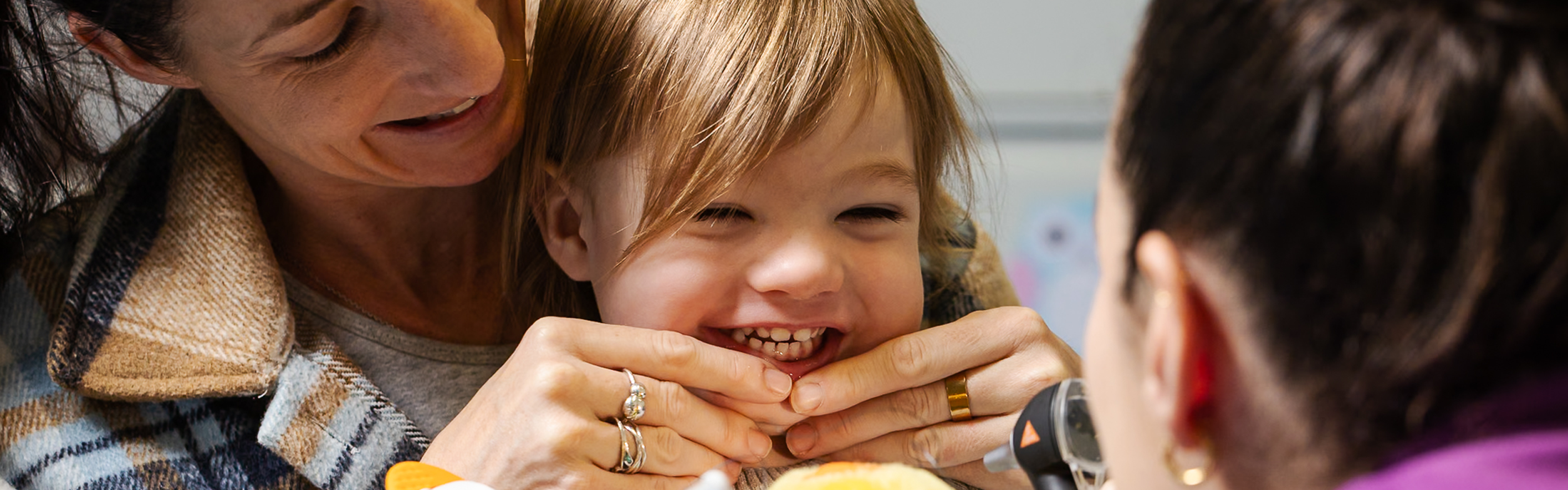 A smiling mum holds her smiling toddler's bottom lip while a dentist prepares to look inside the toddler's mouth