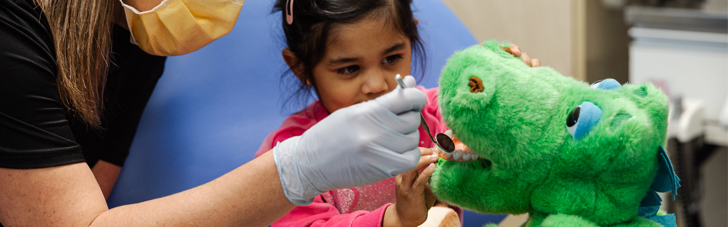 A young girl and dentist checking a toy dinosaur's teeth using a dental mirror